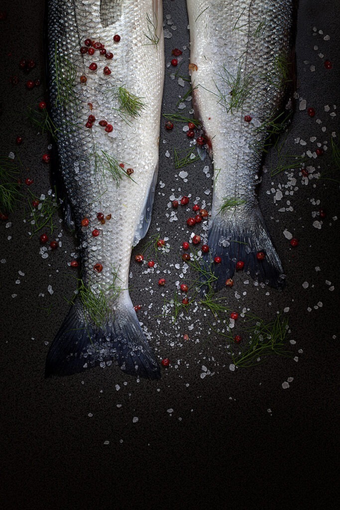 Raw seabass tails with coarse salt, red peppercorn and fresh dill, on black background, top view shot.