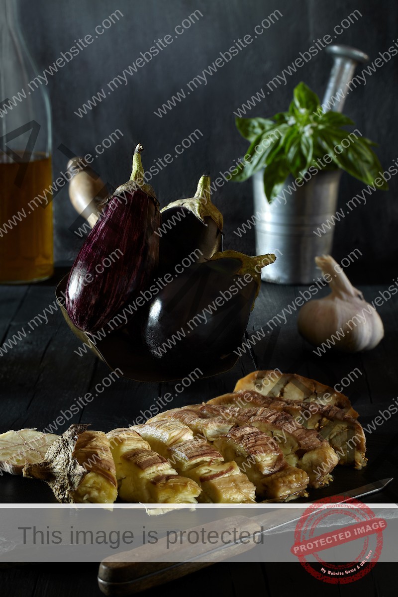 Baked eggplant on slate with raw eggplant in the background.