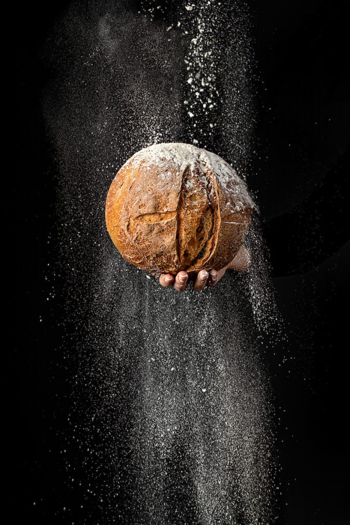 Mano con pagnotta rustica e farina che cade dall’alto su sfondo nero.
