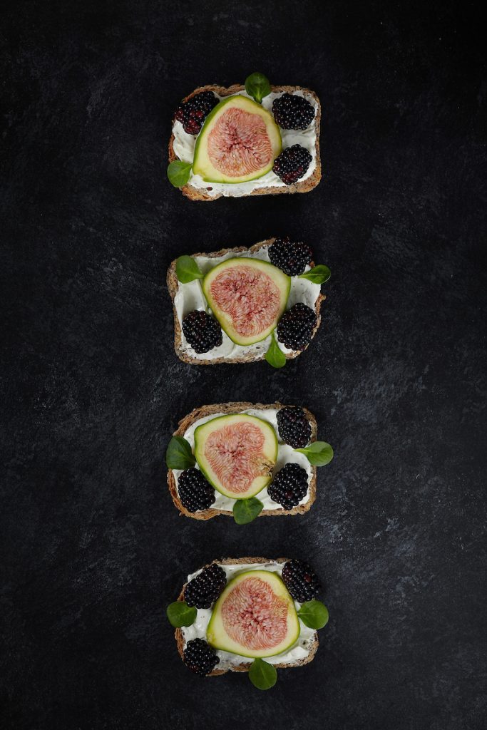 Pane con formaggio spalmabile, fichi, more e valeriana su sfondo scuro.